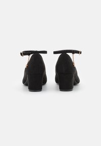 Zapatos de tacón de ante negro con un tacón grueso, tira al tobillo y un detalle decorativo de cadena dorada. Textura suave y diseño de punta redondeada.