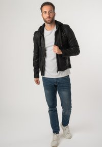 Giacca di pelle nera con cappuccio, chiusura con zip e design testurizzato, indossata sopra una maglietta bianca e jeans blu, abbinata a sneakers bianche.