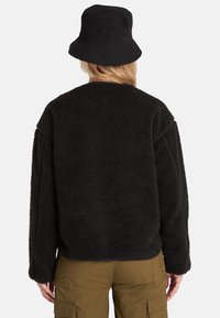 Czarny sweter z polaru o fakturowanej, puszystej powierzchni, luźnym kroju, opadających ramionach i zaokrąglonym dekolcie, zestawiony z czarnym kapeluszem typu bucket.