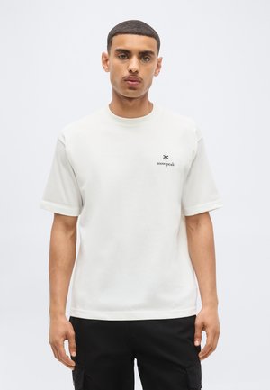 Jeune homme portant un t-shirt blanc uni Snow Peak et un pantalon noir, debout devant un fond clair uni.
