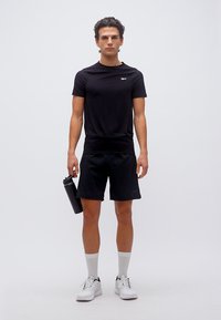 Νεαρός άνδρας με μαύρα αθλητικά ρούχα κρατώντας παγούρι νερού, φοράει λευκά αθλητικά παπούτσια και στέκεται μπροστά σε λευκό απλό φόντο.