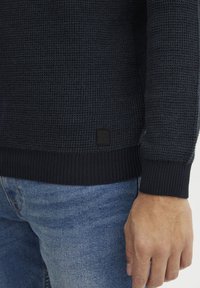 Mørk, strukturert strikkegenser med ribbestrikkede mansjetter og kant; har et lite svart logo-merke på bunnen. Sammen med lyse blå denimjeans.