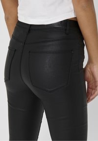 Personne portant un pantalon noir moulant effet cuir avec des poches arrière et un top blanc court visible à la taille.