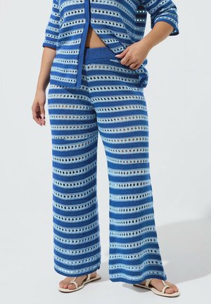 Donna che indossa pantaloni a maglia a righe blu e cardigan abbottonato coordinato, in piedi a piedi nudi con una mano che sistema il cardigan.
