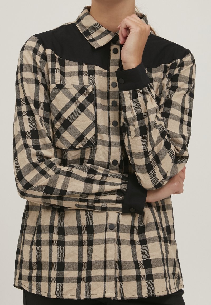 Rutigt skjorta med ett beige och svart rutmönster. Har en solid svart ok, bröstficka och tryckknappar framtill och vid ärmsluten.