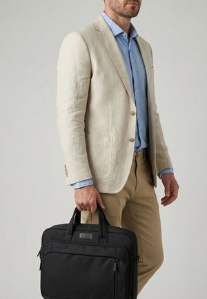 Mand iført beige blazer og khaki bukser, holder sort kufferttaske mod ensfarvet grå baggrund.