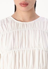 Top bianco, leggero, realizzato in tessuto morbido. Presenta una texture arricciata con plissettature orizzontali e un collo rotondo.