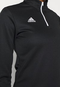 Haut de sport Adidas noir à manches longues avec une fermeture éclair blanche au quart et le logo Adidas sur la poitrine, porté par une personne visible des épaules jusqu'en bas.