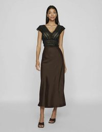 Nő fekete csipkés, rövid ujjú, mély V-nyakú felsőben, sötétbarna szatén, derékban magasított midi szoknyában, fekete magassarkú cipővel.