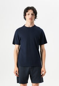 Tricou bleu marin cu mânecă scurtă, din bumbac, cu decolteu rotund. Țesătura pare netedă și este purtat împreună cu pantaloni scurți închiși la culoare.
