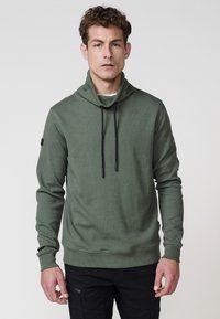 Hombre vistiendo un pulóver verde oliva con cuello alto y cordones negros, combinado con pantalones cargo negros, de pie frente a un fondo plano.