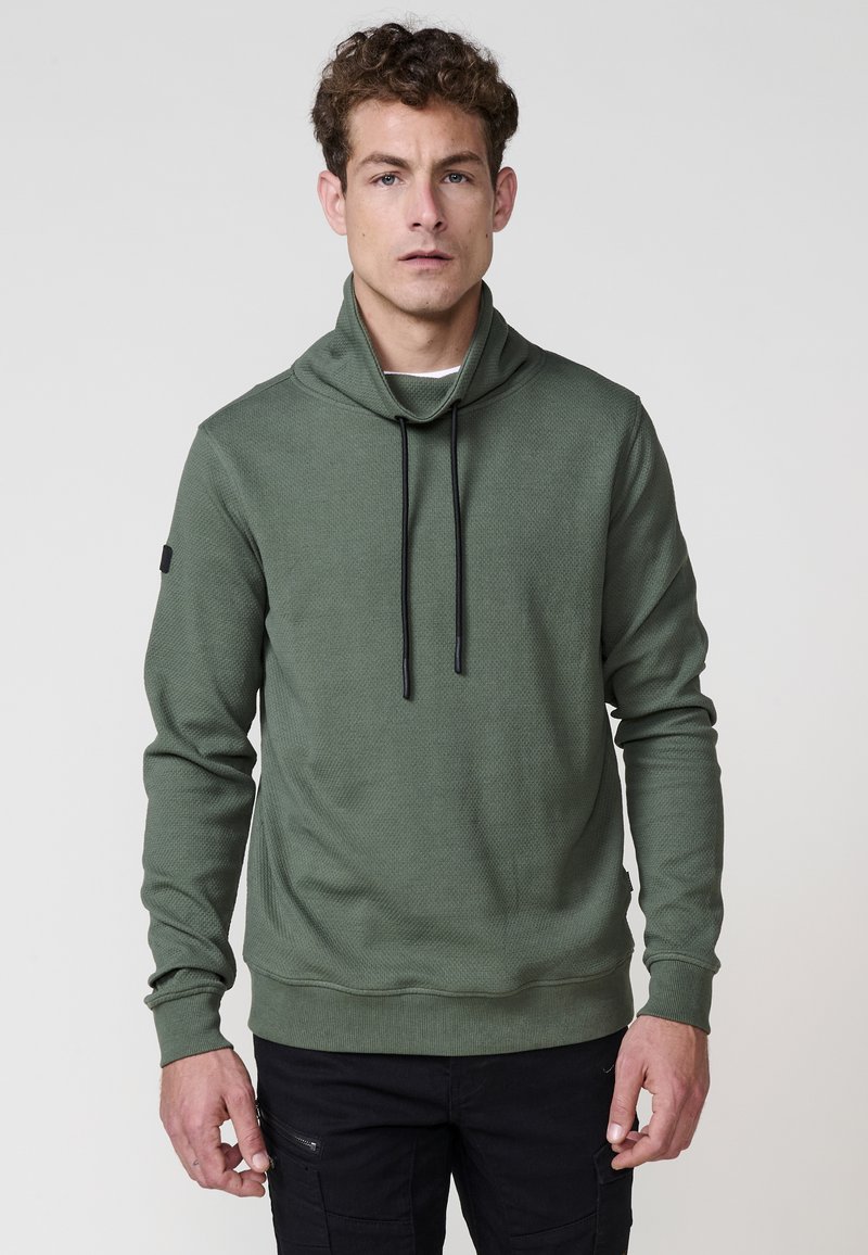 Hombre vistiendo un pulóver verde oliva con cuello alto y cordones negros, combinado con pantalones cargo negros, de pie frente a un fondo plano.