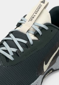 Μαύρο αθλητικό παπούτσι με υφή, ανοιχτομπλε κορδόνια, γλώσσα σε κρεμ απόχρωση, ριγωτό υλικό και μπεζ λογότυπο Nike swoosh. Ετικέτα "Juniper Trail" στη γλώσσα.