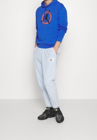 Blauer Pullover-Hoodie mit schwarzem und orangefarbenem Logo, kombiniert mit hellblauen Jogginghosen mit einem weißen Logopatch und schwarzen Sneakers mit grünen Akzenten.