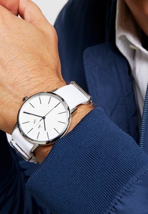Gros plan sur un poignet portant une montre analogique argentée avec un cadran blanc, des marqueurs d'heure noirs minimalistes et un bracelet en tissu blanc.
