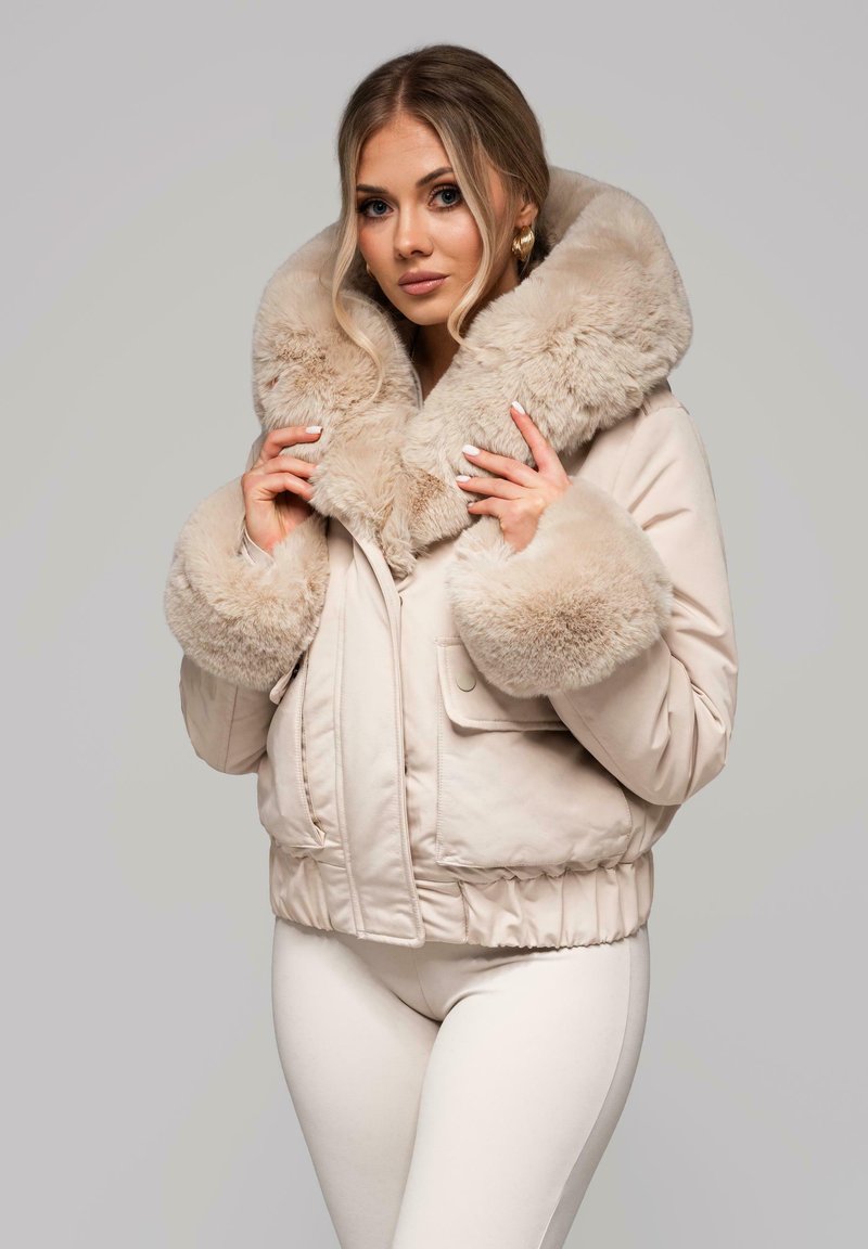 Jasnobeżowa kurtka typu bomber z dużym, miękkim kołnierzem i mankietami z sztucznego futra. Posiada przednie kieszenie oraz elastyczny pas w talii.