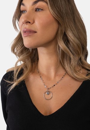 Frau mit welligem blondem Haar, die ein schwarzes V-Ausschnitt-Oberteil und eine silberne Halskette trägt, die einen runden Anhänger mit einem kleinen Sonnenanhänger darin zeigt.