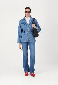 Jeansjacke mit Gürtel, hellblaue Farbe, vorne mit Knöpfen, zwei Brusttaschen. Kombiniert mit hellblauen Schlagjeans und roten Schuhen. Schwarze Tasche.