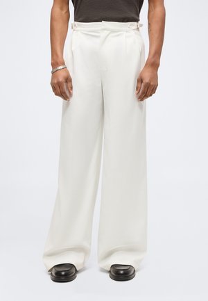 Homme portant un pantalon blanc taille haute à jambes larges avec des boucles latérales, des chaussures noires et un haut à manches courtes foncé, debout devant un fond uni.