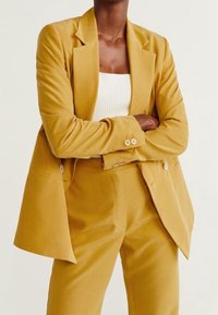 Blazer jaune moutarde à coupe décontractée, à simple boutonnage avec fermeture à deux boutons, associé à un pantalon assorti et un haut blanc.