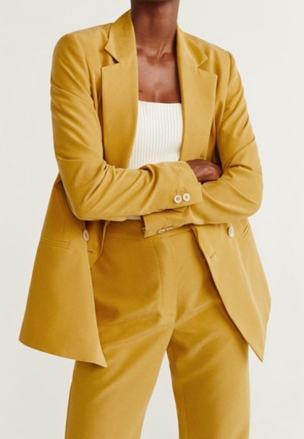Blazer jaune moutarde à coupe décontractée, à simple boutonnage avec fermeture à deux boutons, associé à un pantalon assorti et un haut blanc.