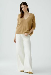 Beige gestrickter Pullover mit V-Ausschnitt und kurzen Ärmeln, kombiniert mit weißen, weiten Hosen. Weiche Textur, entspannte Silhouette, minimale Details.