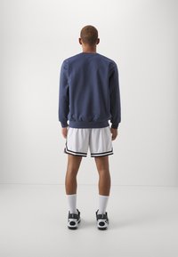 Sweatshirt azul-marinho com punhos canelados, usado com calções brancos com riscas pretas. Ténis pretos e meias brancas completam o visual.