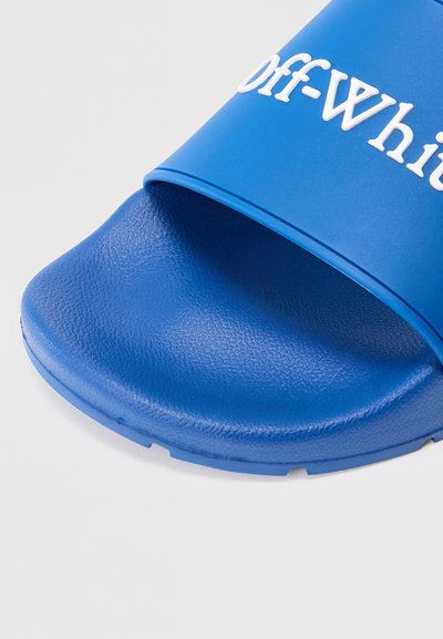 Kék gumi csúszdaszalag, textúrázott felülettel és széles pánttal, amelyen fehér "Off-White" felirat található. Kerek orr és lapos talp.