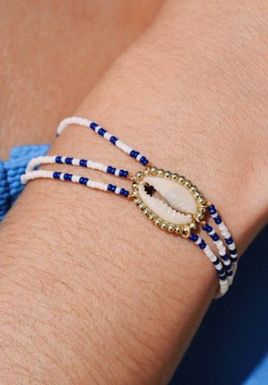 Bracelet en perles avec des perles blanches et bleues, des accents dorés et une coquille de coquillage centrale. Porté sur un poignet avec un teint de peau bronzé.