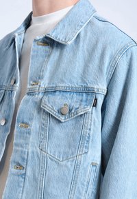 Chaqueta de denim azul claro con un diseño clásico, que presenta botones plateados, dos bolsillos en el pecho y un cuello ligeramente deshilachado.