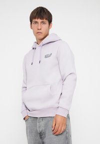 Lavender hoodie made of soft fabric, featuring a front pocket, drawstring hood, and small logo detail on the chest, paired with light grey jeans.