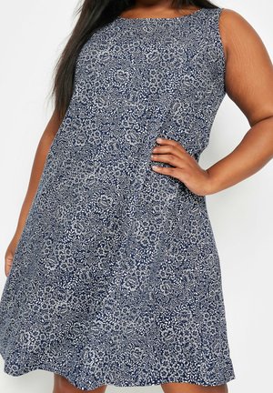 Femme portant une robe bleu marine sans manches avec imprimé floral blanc, main sur la hanche, sur fond blanc uni.