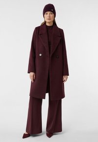 Cappotto in misto lana color bordeaux con design doppiopetto, grandi rever e due bottoni frontali; abbinato a pantaloni a gamba larga coordinati e un berretto.