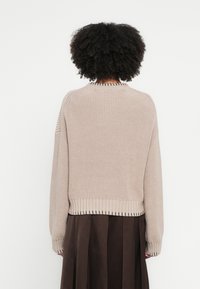 Bēša adīta džemperis ar ribotu apakšu un aprocēm, kuram ir teksturēts raksts un apaļa kakla izgriezums. No aizmugures redzams brīvs piegriezums.