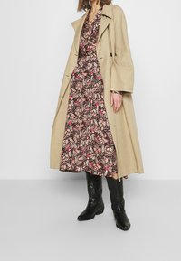 Cappotto trench beige su un vestito marrone a fiori. Stivali neri fino al ginocchio con tacco basso. Tessuto morbido e silhouette fluida.