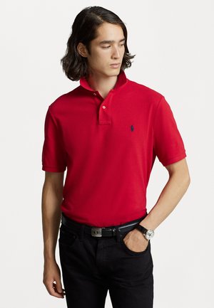 Rode polo shirt met een omslagkraag, twee knopen en een klein blauw logo op de borst, gecombineerd met een zwarte broek en een horloge.