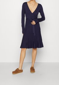 Mörkblå omslagsklänning med långa ärmar, plisserad kjol och ett band i midjan. Texturerad tyg och vågig nederkant. Bruna halkskyddsskor.