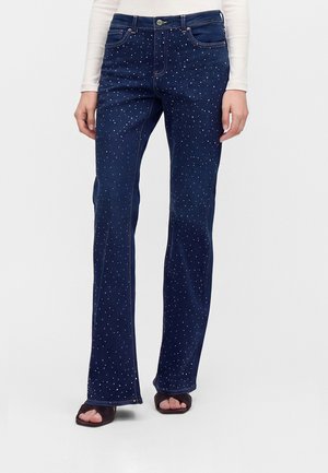 Jean évasé bleu foncé à petits pois blancs, porté avec des sandales noires à bout ouvert et un haut blanc à manches longues.