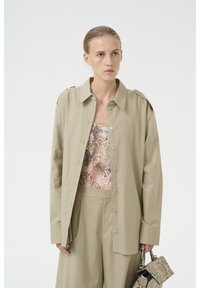 Svijetlo khaki oversized košulja s prednjim gumbima, dugim rukavima i epauletima na ramenima, uparen s teksturiranim topom sa zmijskim uzorkom i hlačama.