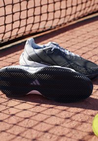 Sort-hvide tennissko med teksturerede såler på en rød grus tennisbane nær nettet og en tennisbold.