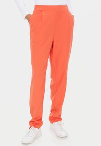 Korallenorange Hosen mit glatter Textur, schmaler Passform, Seitentaschen und Vorderfalten, kombiniert mit weißen Sneakers.