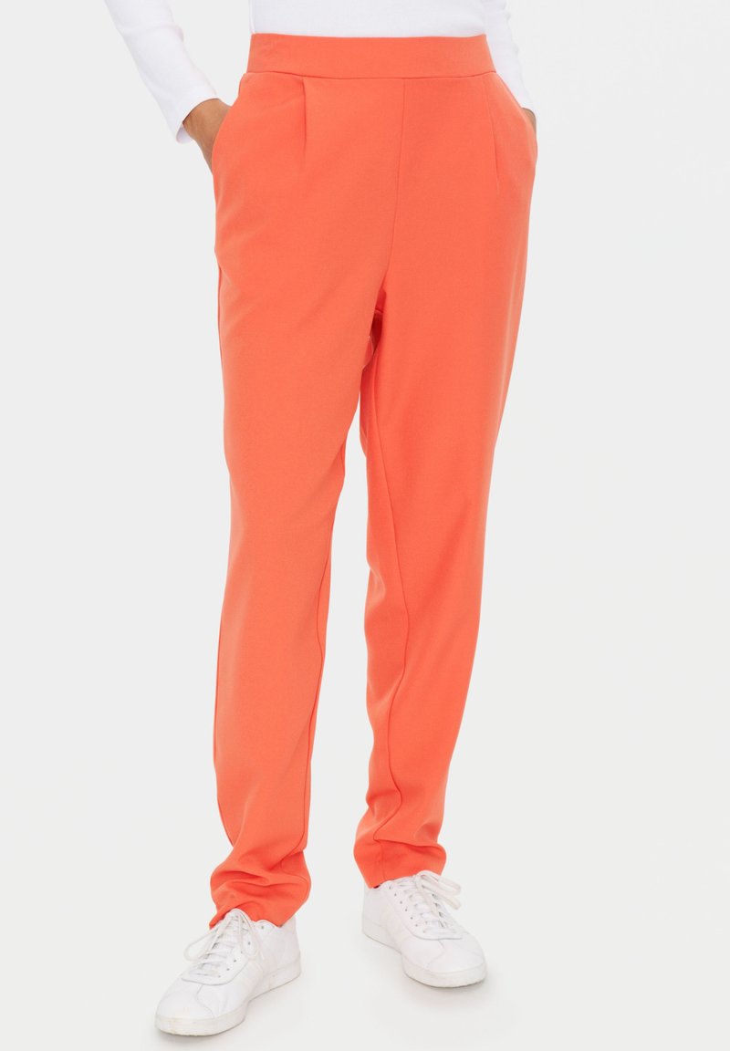 Korallenorange Hosen mit glatter Textur, schmaler Passform, Seitentaschen und Vorderfalten, kombiniert mit weißen Sneakers.