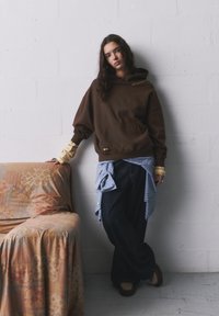 Hnedá oversized mikina s predným vreckom a zlatým textom. Nosená cez svetlo modrú košeľu a tmavé džínsy. V blízkosti textúrovaná, vzorovaná pohovka.