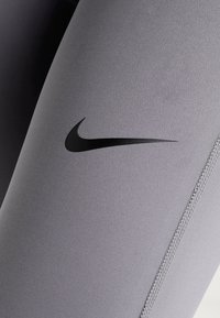 Grå strækleggings med en glat tekstur, der har et fremtrædende sort Nike-logo på låret og kontrasterende syninger langs sømmene.