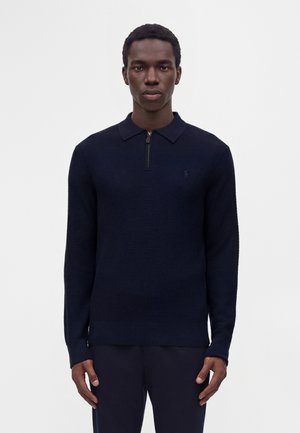 Jeune homme portant un pull zippé texturé bleu marine foncé et un pantalon assorti, debout devant un fond clair uni.