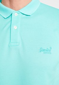 Polo shirt turquoise clair en tissu texturé, avec un col, deux boutons blancs et un logo brodé sur le côté gauche.