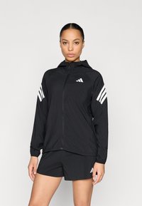 Chaqueta cortavientos negra con capucha, con detalles de tres franjas blancas en las mangas y logo en el pecho, fabricada con un material ligero.