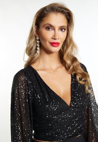 Zwarte pailletten top met lange, doorschijnende mouwen en een diepe V-hals. Model draagt een opvallende rode lippenstift en hangende versierde oorbellen.