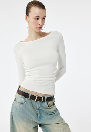 Junge Frau trägt ein weißes, enganliegendes Langarmshirt, große Creolen und lockere hellblaue Jeans mit schwarzem Gürtel, schaut zur Seite.