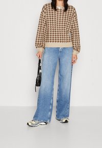 Beige och brun hundtandsmönstrad tröja med puffärmar, tillsammans med ljusblå vida jeans och en svart väska. Sportiga flerfärgade sneakers.